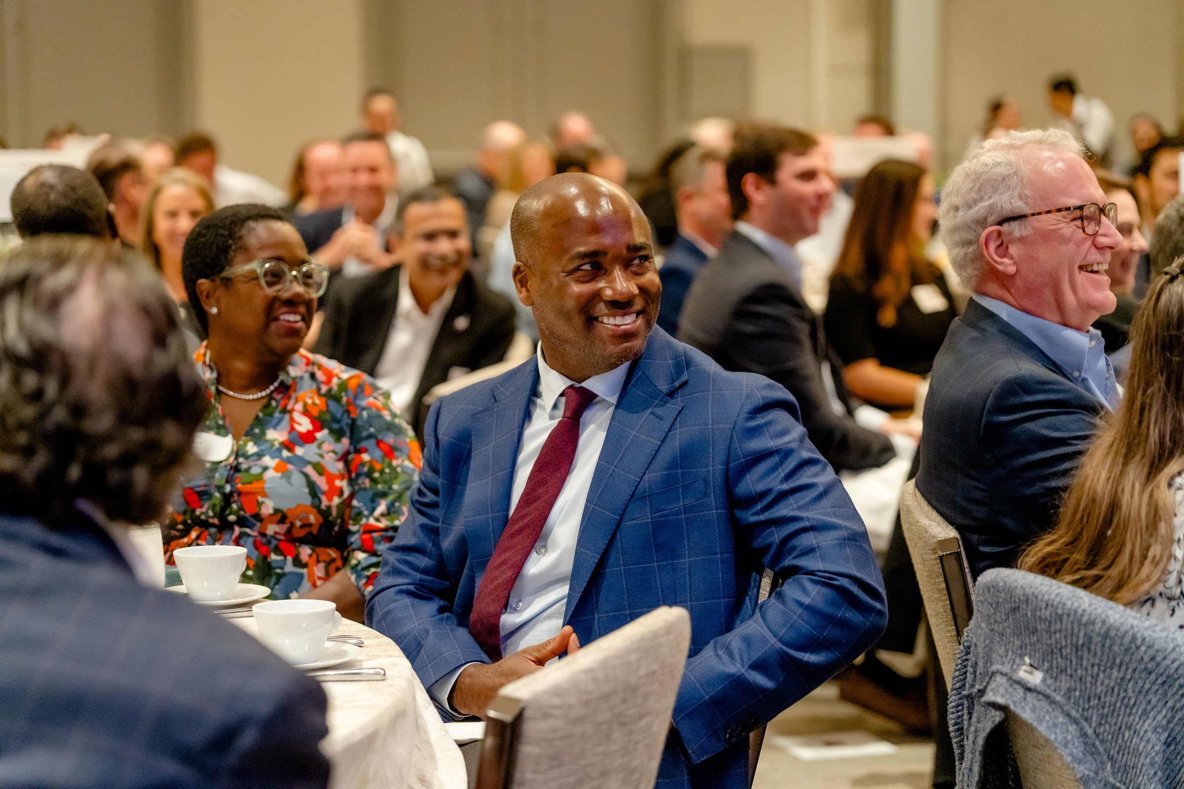 Guests at a luncheon table smiling toward the stage, a full room behind them